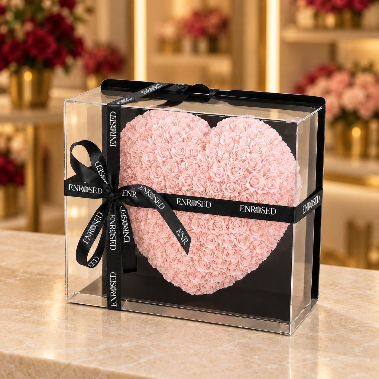 Pink foam rose heart (25 cm) in a clear box with black ENROSED ribbon, on a beige showroom counter.